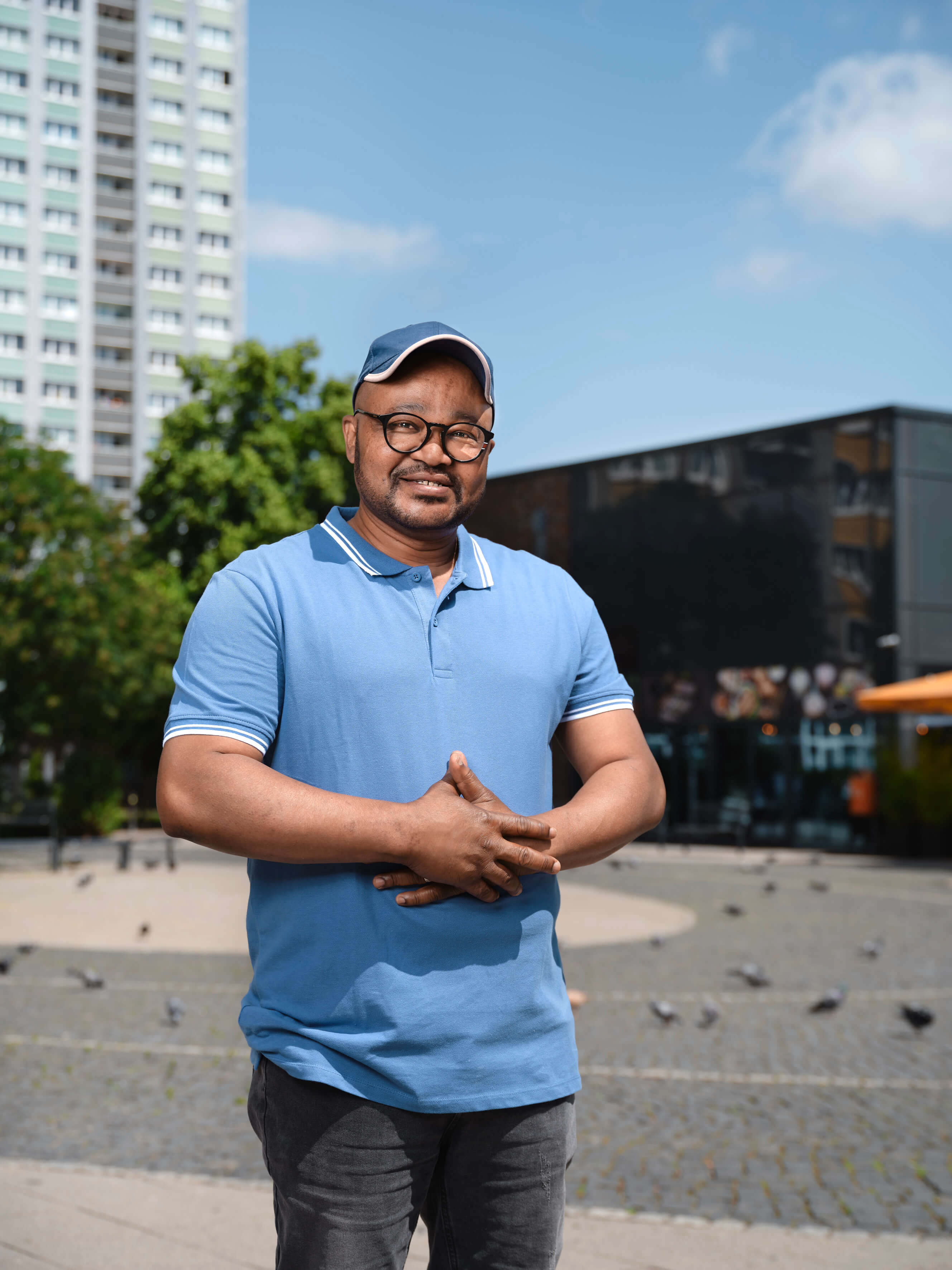 Eine Person mit schwarzer Brille, blauen Cap, blauen Polo-Shirt und schwarzer Hose steht vor einem Hochhaus und lächelt in die Kamera