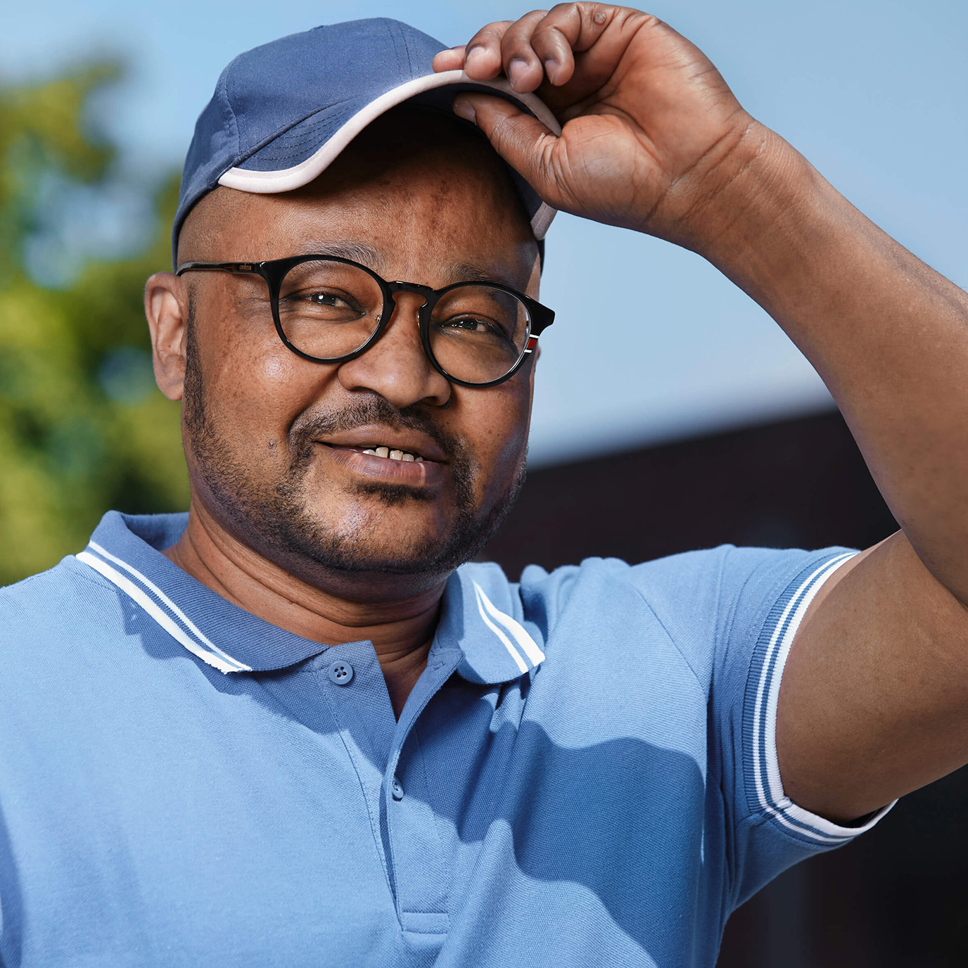 Eine Person mit schwarzer Brille, blauen Cap und blauen Polo-Shirt steht vauf einer Straße. Hinter ihm sieht man grüne Bäume und ein braunes Haus.