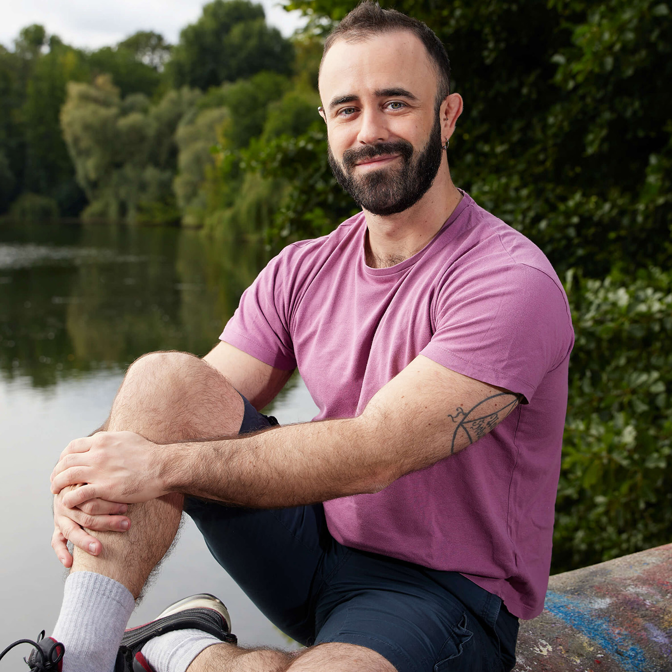 Eine Person mit kurzen dunklen Haaren und Bart, rosa Shirt und dunklen Shorts sitzt auf einer Mauer vor einem Waldsee und lächelt in die Kamera.