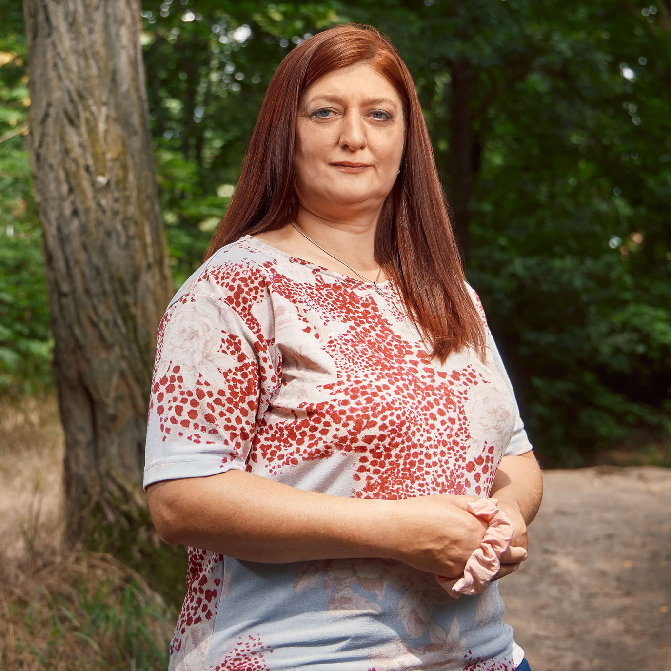 Eine Person mit langen roten Haaren, hellblau-rot gemusterten Shirt und dunklen Jeans steht vor einem Waldweg und lacht in die Kamera.