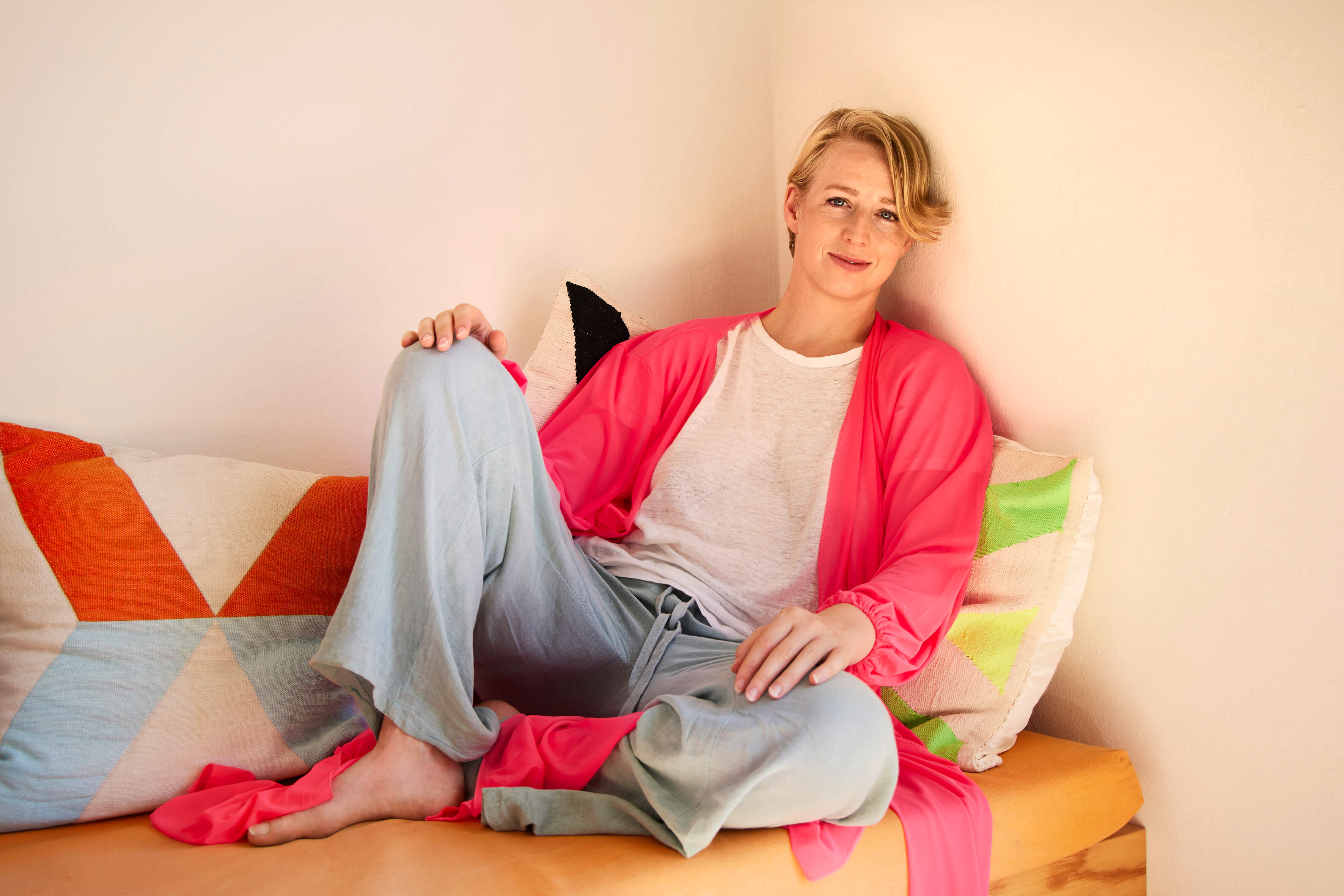 Eine Person mit kurzen blonden Haaren, weißem T-Shirt und langem rosa Cardigan sowie hellblauen Jeans sitzt auf einem Sofa mit bunten Kissen und lächelt in die Kamera.