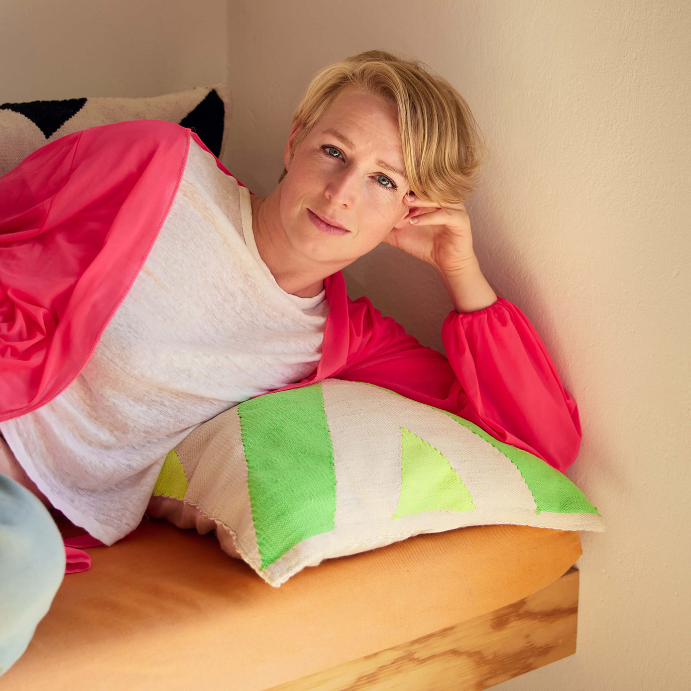 Eine Person mit kurzen blonden Haaren, weißem T-Shirt und langem rosa Cardigan sowie hellblauen Jeans sitzt auf einem Sofa mit bunten Kissen und lächelt in die Kamera.