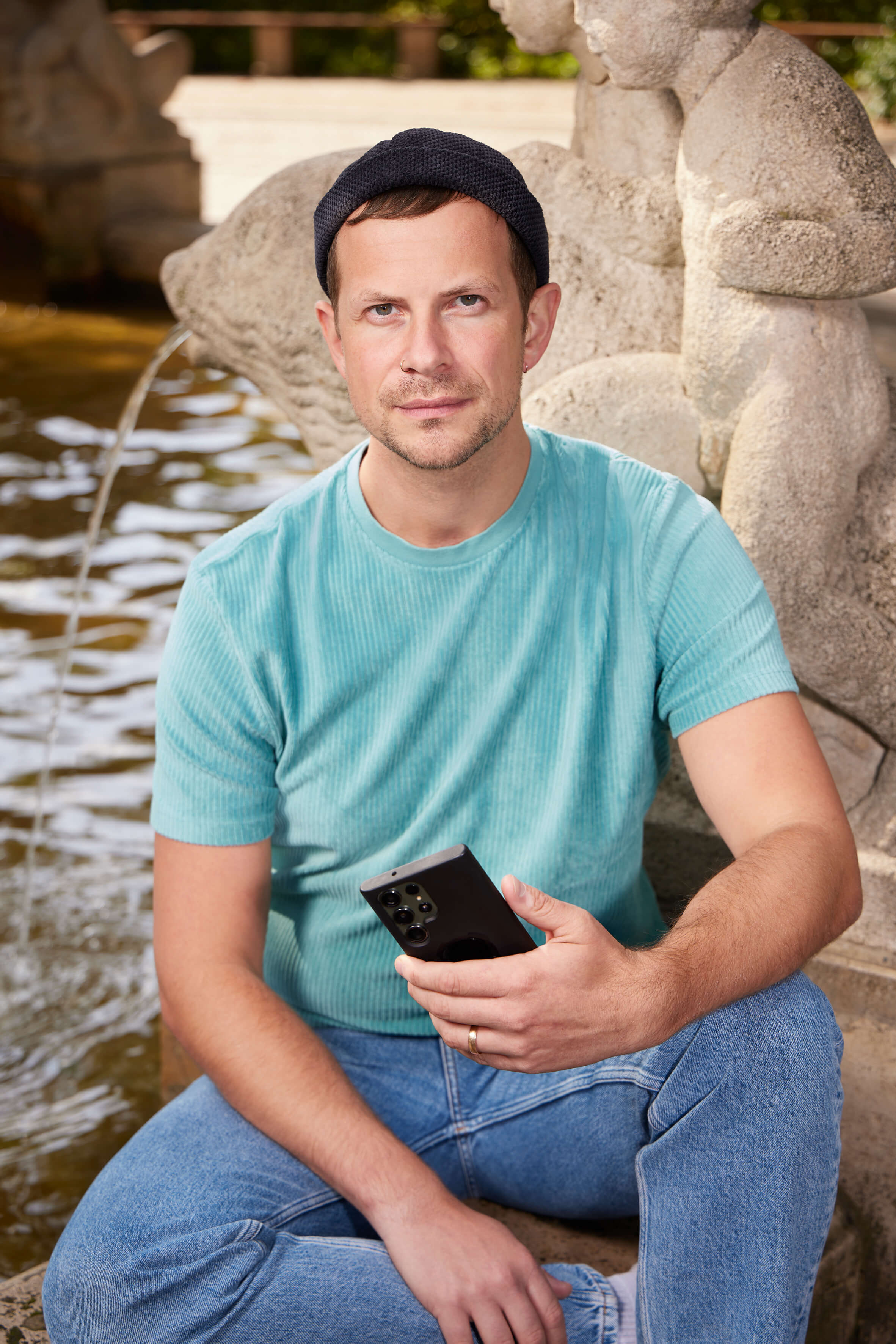 Eine Person mit kurzen dunklen Haaren, dunkelblauer Mütze und hellblauem T-Shirt sitzt mit einem Handy in der Hand an einem Stadtbrunnen und schaut in die Kamera. 