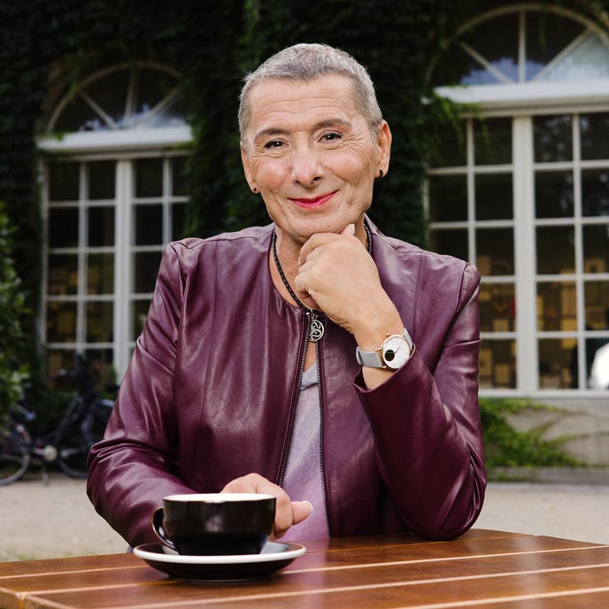 Eine Person mit ganz kurzen grauen Haaren und violetter Lederjacke sitzt mit einem Kaffee am Gartentisch und lächelt in die Kamera.