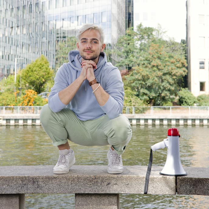 Eine Person mit blonden Haaren und dunklen Bart, hellblauen Hoodie sowie grüner Hose sitzt vor einem Glasgebäude und lächelt in die Kamera.