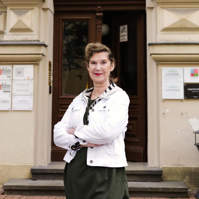 Eine Person mit dunklem Pferdeschwanz, schwarzem Kleid und weißer Jeansjacke steht mit verschränkten Armen vor einem Eingang eines Altbaus und lächelt in die Kamera.